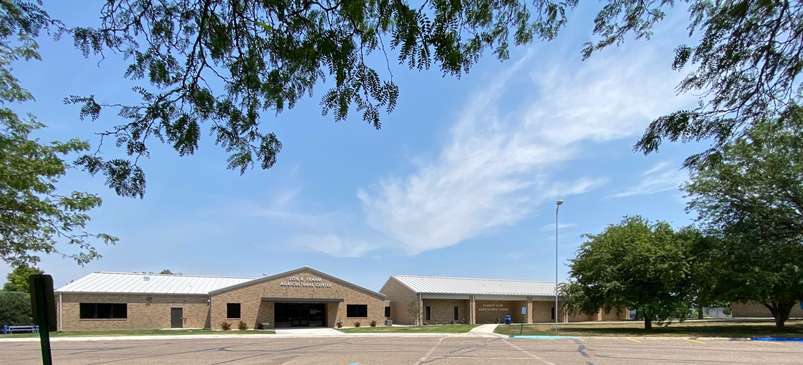 The Lon R. Frahm and Stanley Carr Agricultural Centers, tan brick buildings with white roofs, sit side by side. Tree branches frame the top, and the sky is clear with light clouds.